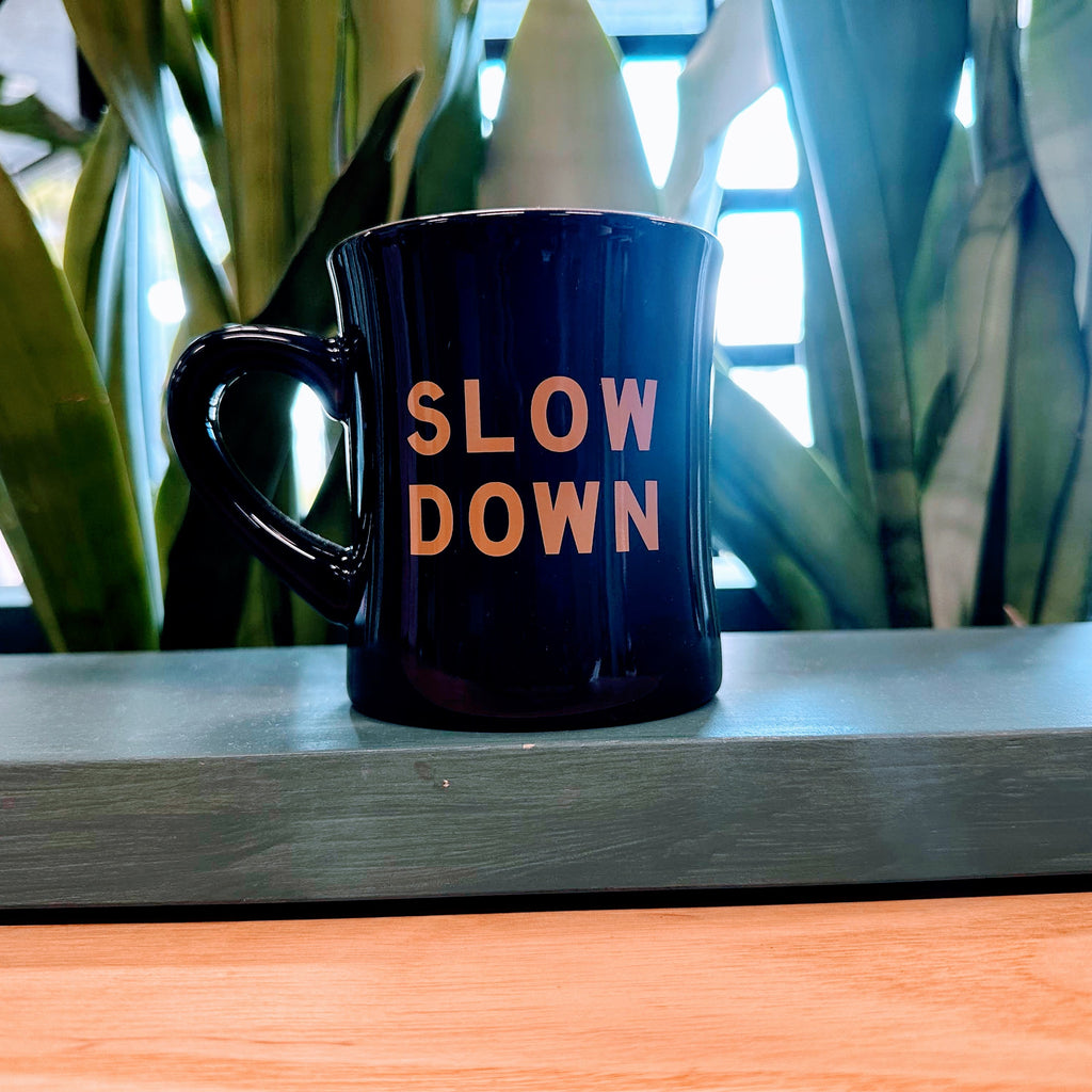 Black mug with 'SLOW DOWN' text on a wooden surface with plants in the background