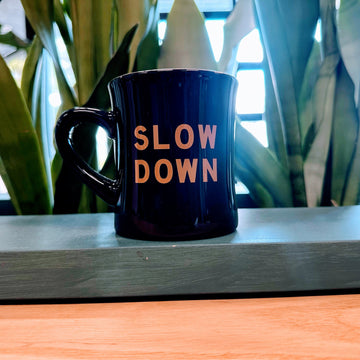 Black mug with 'SLOW DOWN' text on a wooden surface with plants in the background